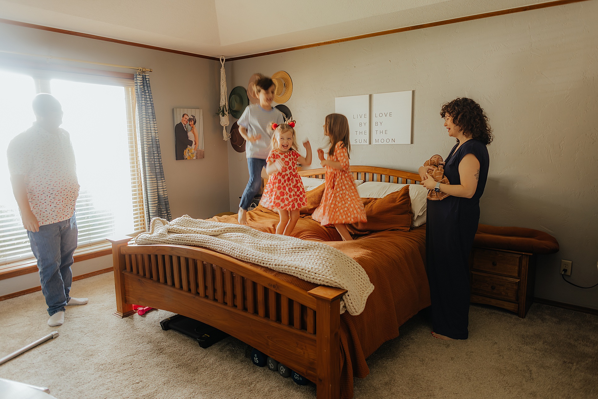 mom holds newborn as older siblings jump on bed