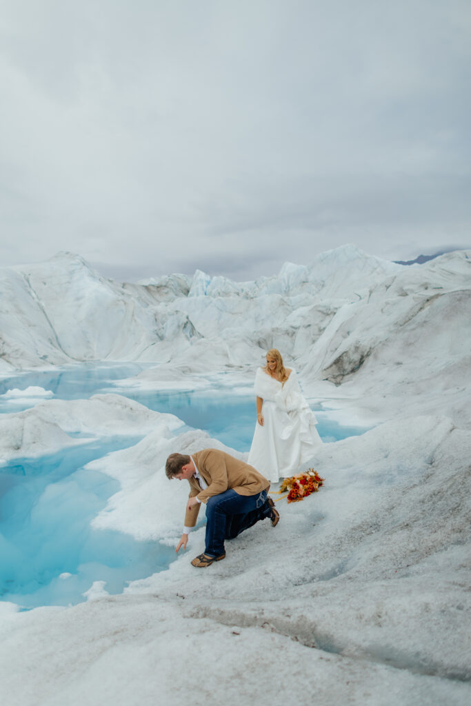 Groom touching the blue glacial ice while the bride stands beside a melt pool on Knik Glacier in Alaska.