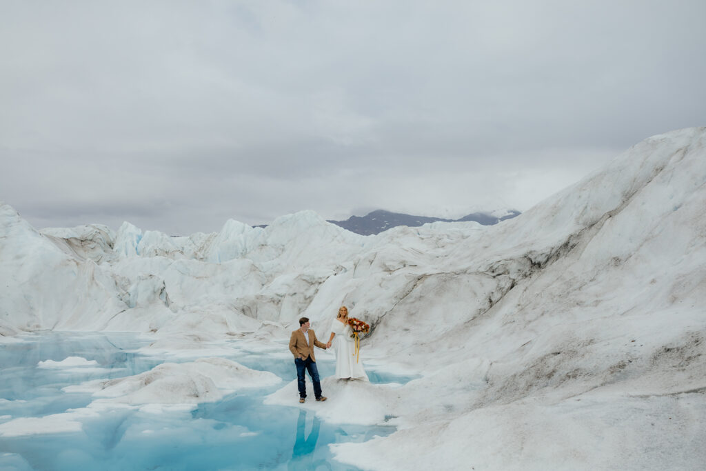 Bride and groom standing together on Knik Glacier surrounded by bright blue melt pools and ice formations.