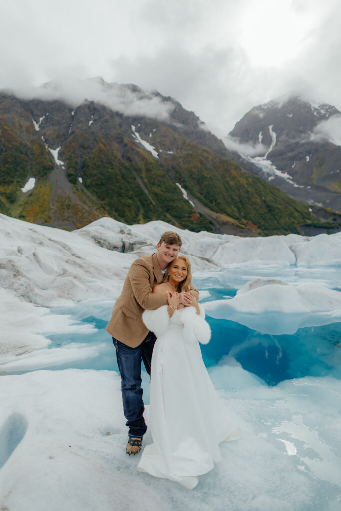Newlyweds smiling together on Knik Glacier with turquoise melt pools behind them.