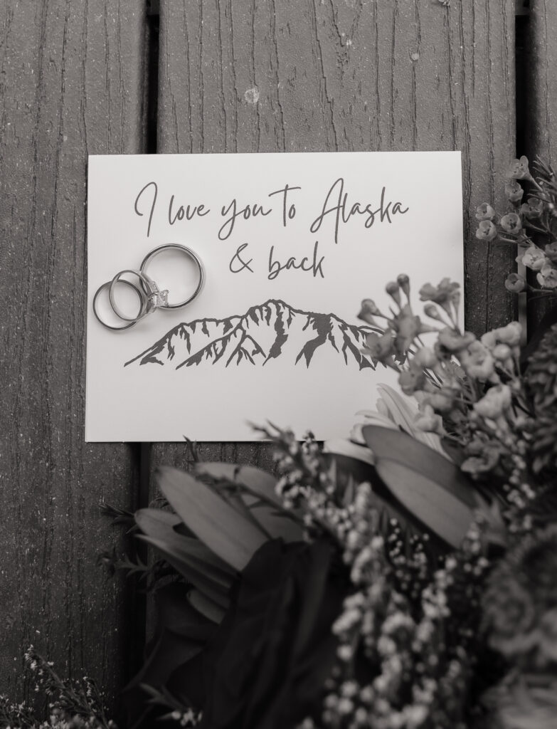 Wedding rings resting on an "I love you to Alaska and back" card with bouquet flowers before a Knik Glacier helicopter elopement in Alaska.